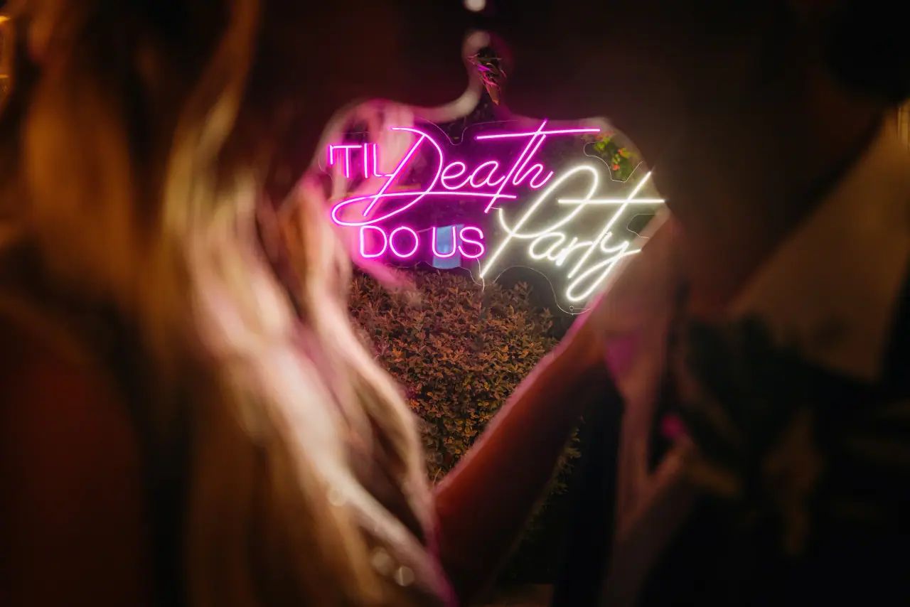 Neon sign reading 'Til Death Do Us Party' glowing beyond a blurred guest