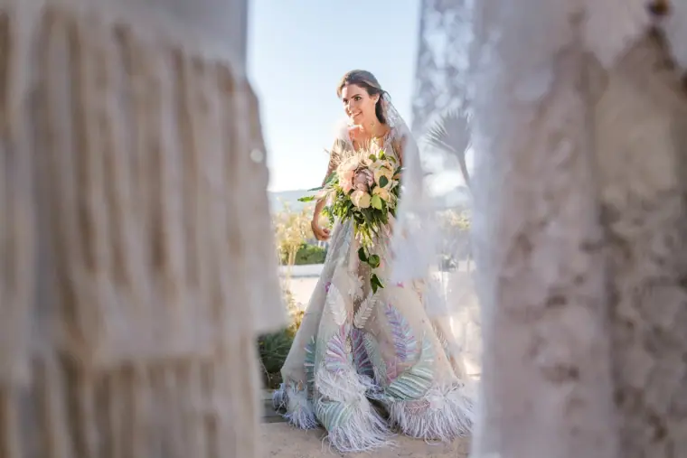 Hassiba in her feathered gown and veil holding her bouquet, framed by macramé