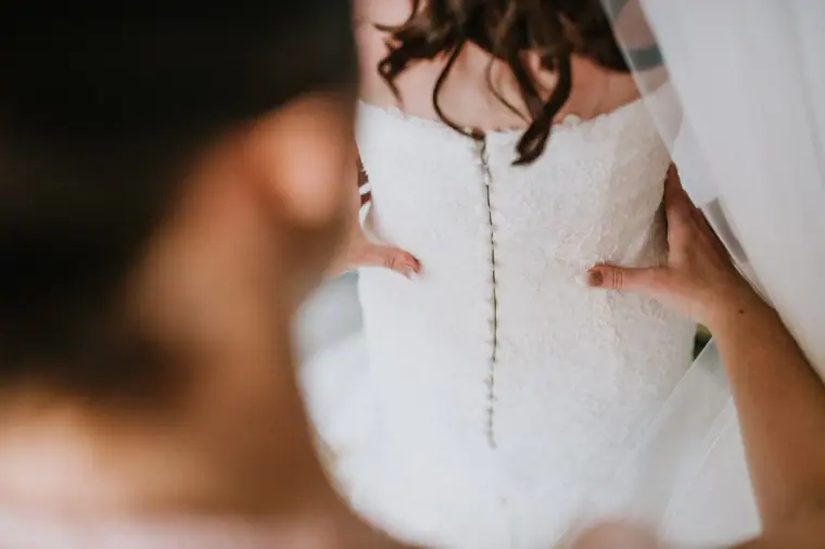 Bridesmaid buttoning the back of Claire's lace wedding dress