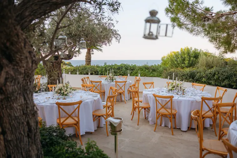 Reception tables with wooden cross-back chairs beneath olive trees by the sea
