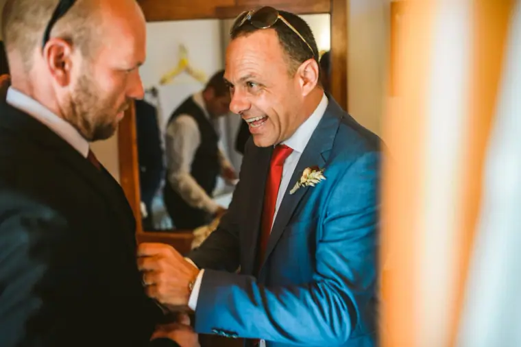 Man in a blue suit laughing as he helps the groom with his buttonhole