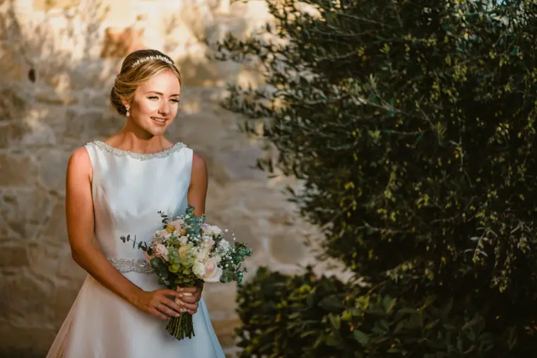 Nicola in a high-neck gown holding her bouquet beside an olive tree at Liopetro