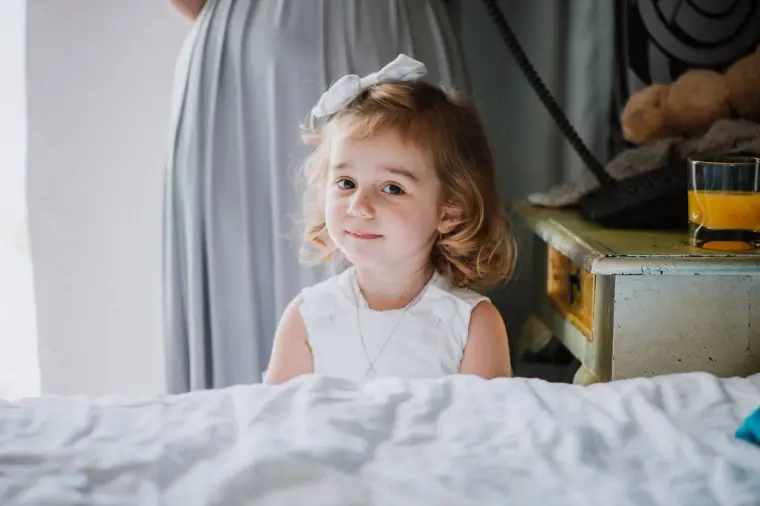 Little flower girl in a white dress peeking over the bed