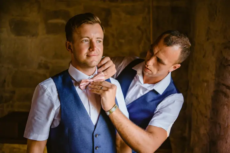 Best man fastening the groom's pink bow tie in a stone room