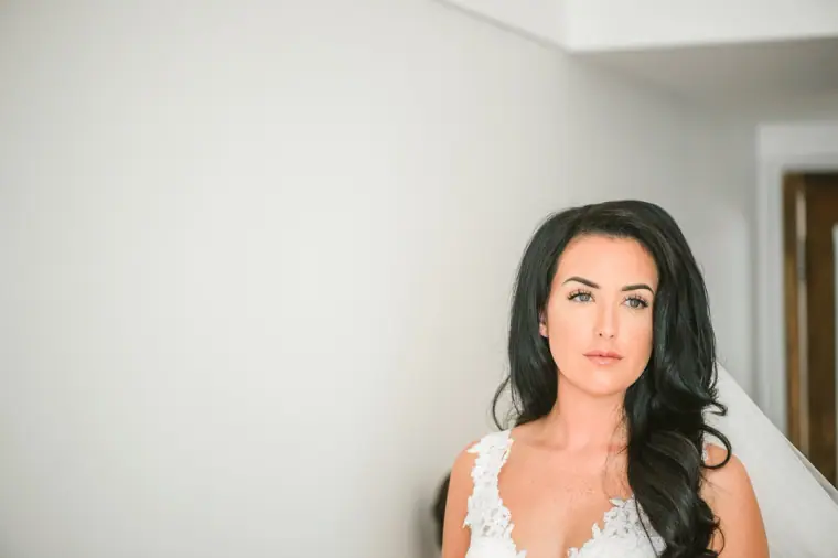 Portrait of Jaclyn in her lace wedding gown against a white wall, dark hair over one shoulder