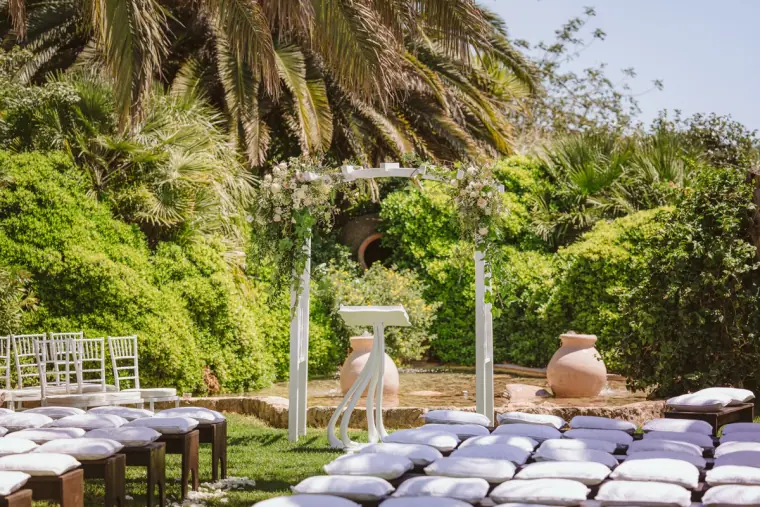 Garden ceremony with white benches, cushions and a floral arch among palms