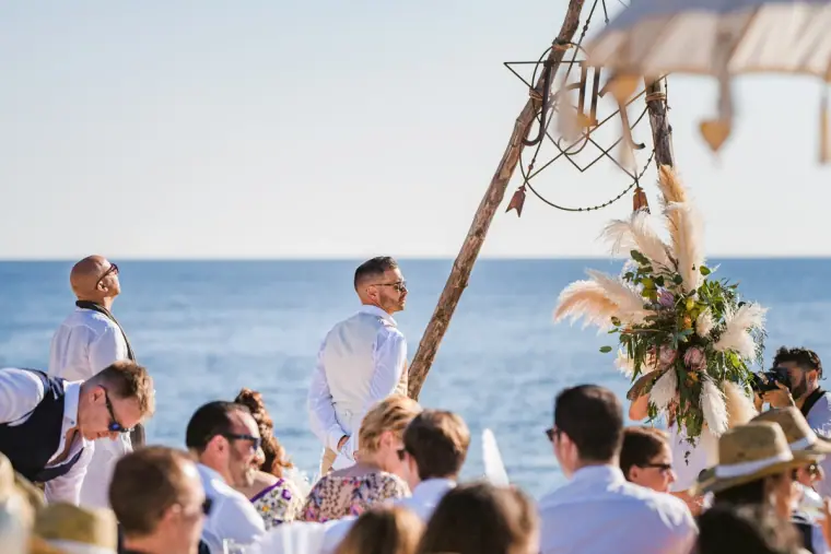 Groom waiting under the tipi arch as guests gather by the sea