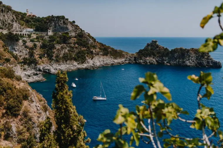 Blue Amalfi Coast cove with sailing boats, cliffs and fig leaves in the foreground