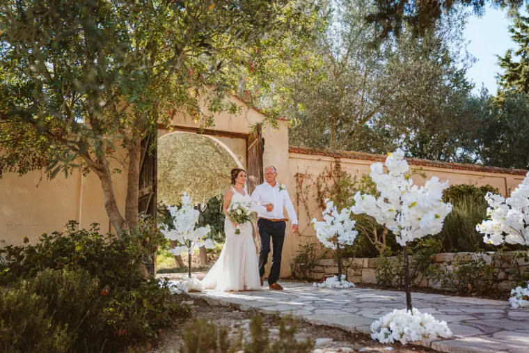 Bride walking into the courtyard on her father's arm past white blossom trees