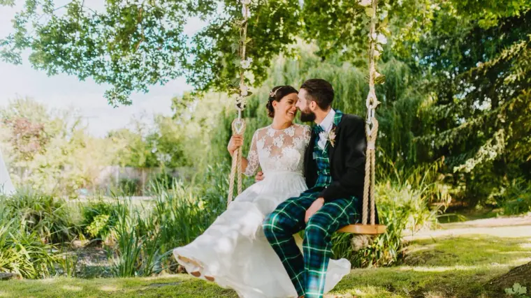 Bride in lace and groom in green tartan trousers laughing together on a garden swing in Windsor