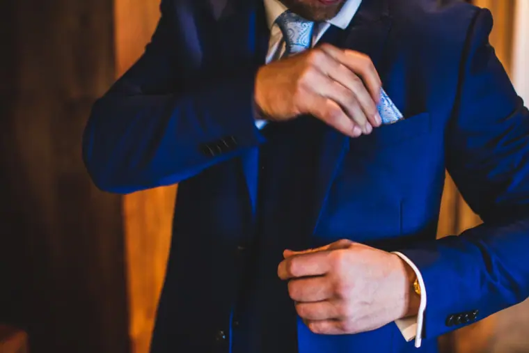 Close-up of the groom tucking a pocket square into his navy suit jacket