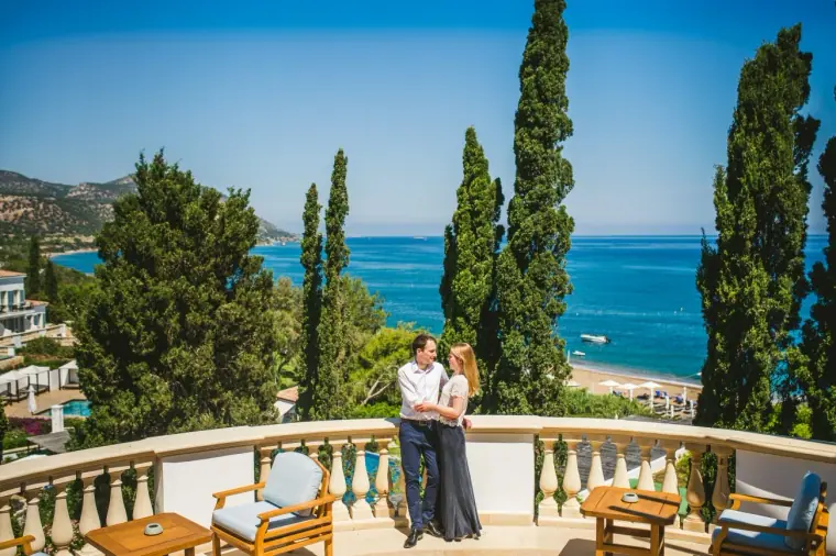 Alison and Daniel on a sunny Anassa terrace with cypresses, armchairs and blue bay beyond, Polis