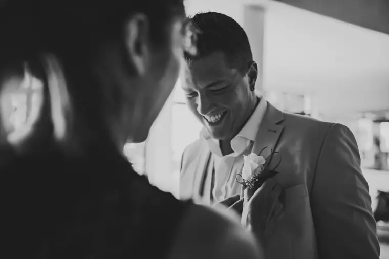 Black-and-white photo of the groom laughing as a buttonhole is pinned to his jacket