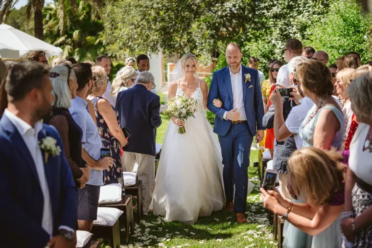 Sophie walking down the garden aisle on a guest's arm as guests turn to watch