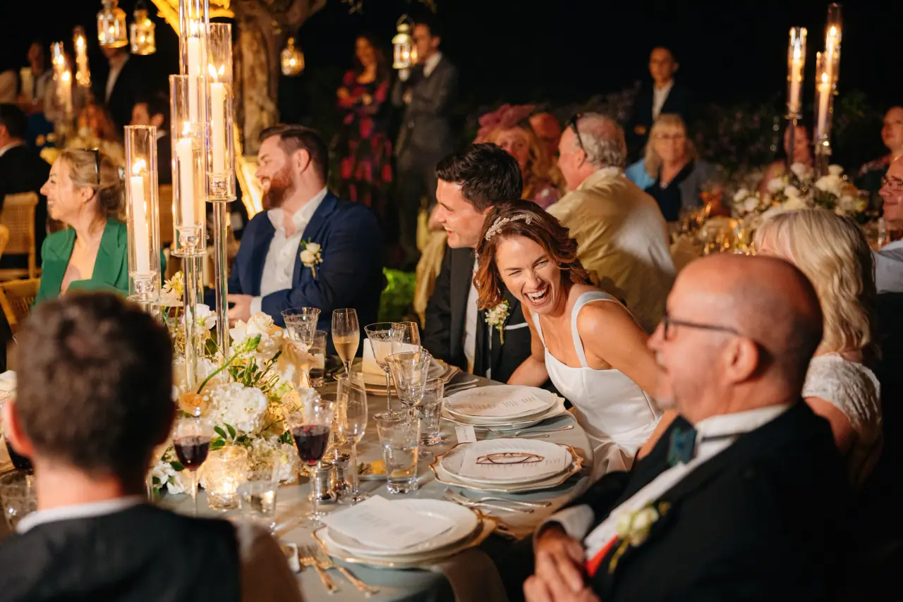 Guests laughing around a candlelit dinner table at Mihail and Georgina's Anassa reception