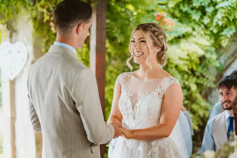 Bride smiling at the groom as they hold hands during the ceremony under the pergola