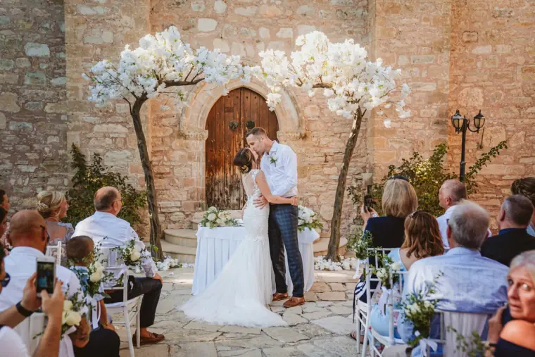 Ellie and Jack kissing beneath white blossom trees by the chapel door as guests watch