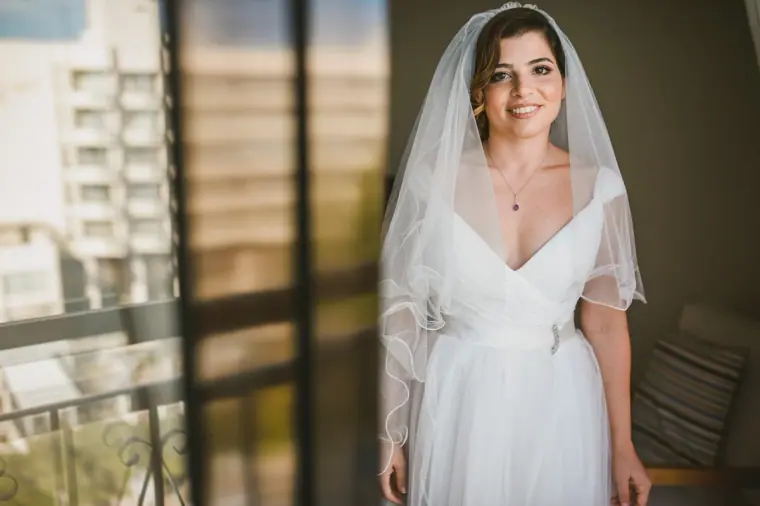 Nermine smiling in her V-neck wedding dress and veil by a balcony door