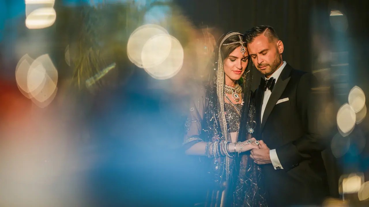 Bride in an embroidered South Asian outfit and groom in a tuxedo amid colourful bokeh
