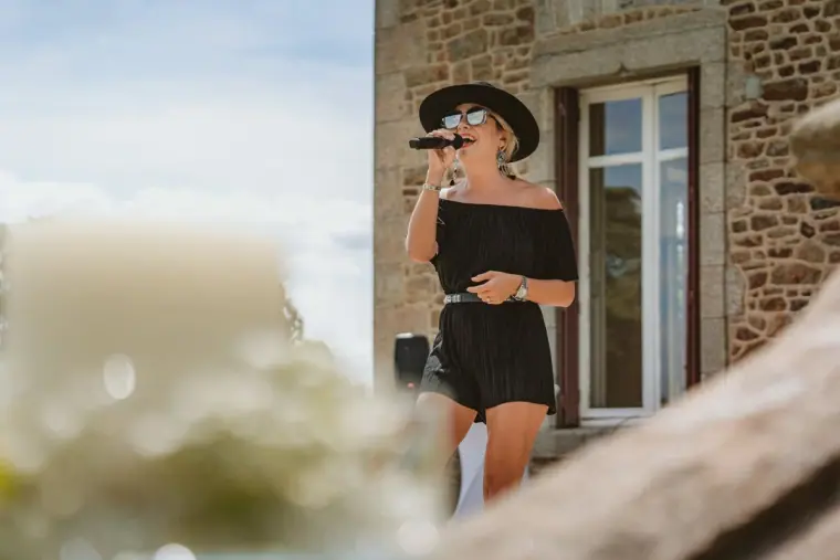 Female guest in a black hat and off-shoulder dress speaking beside the château wall