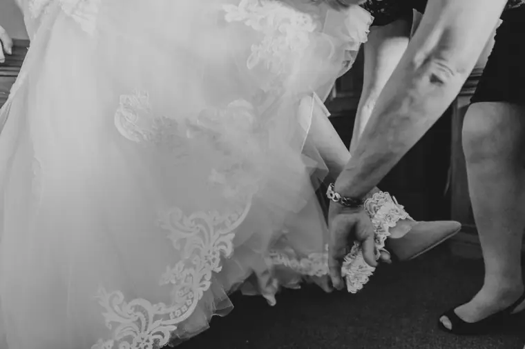 Black-and-white photo of a bride's lace hem as she slips on her shoes