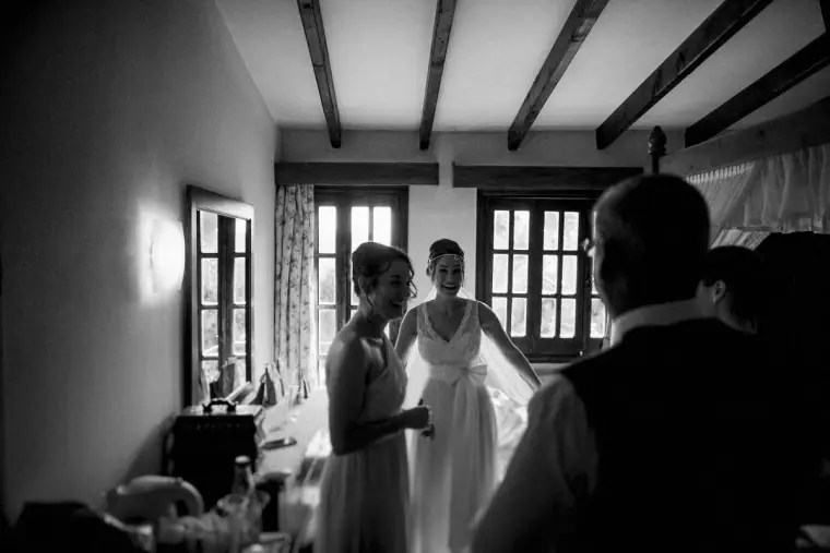 Black-and-white photo of the bride standing in a beamed room as her dress is checked