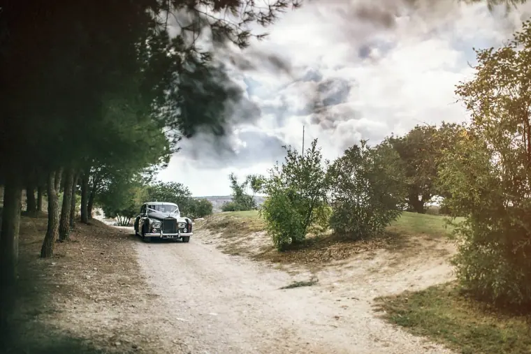 Vintage car driving up a dusty tree-lined track towards the venue