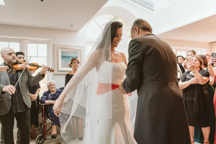 Bride's father lifting her veil as relatives and musicians look on