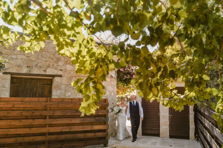 Bride and her father stepping out from a stone archway under leafy branches