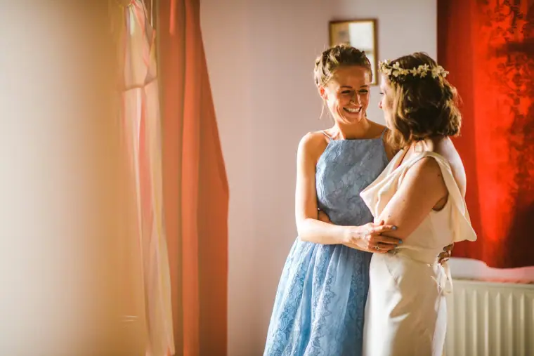 Bride in a flower crown embracing a bridesmaid in blue lace by a red curtain