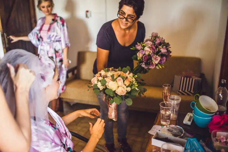 Bride in a floral robe as a friend brings in pastel bouquets