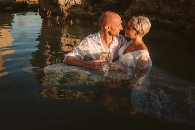 Alana and Luke leaning together on a submerged rock, toasting with champagne
