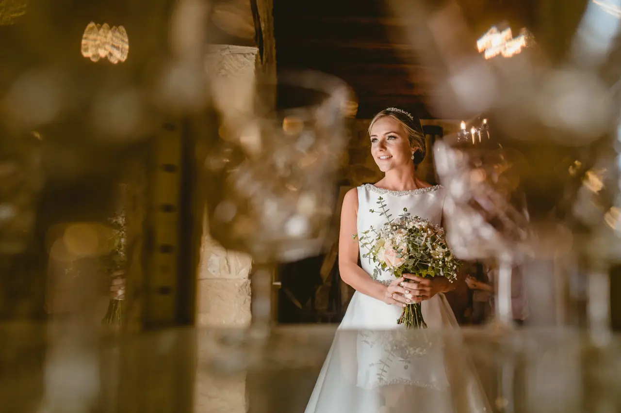 Bride with her bouquet framed by blurred glassware in a rustic room