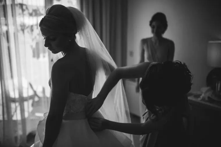 Black-and-white photo of a bridesmaid fastening Charlotte's dress by the window