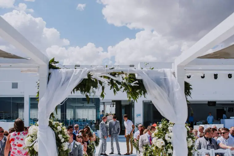 Draped white ceremony arch with guests gathering behind