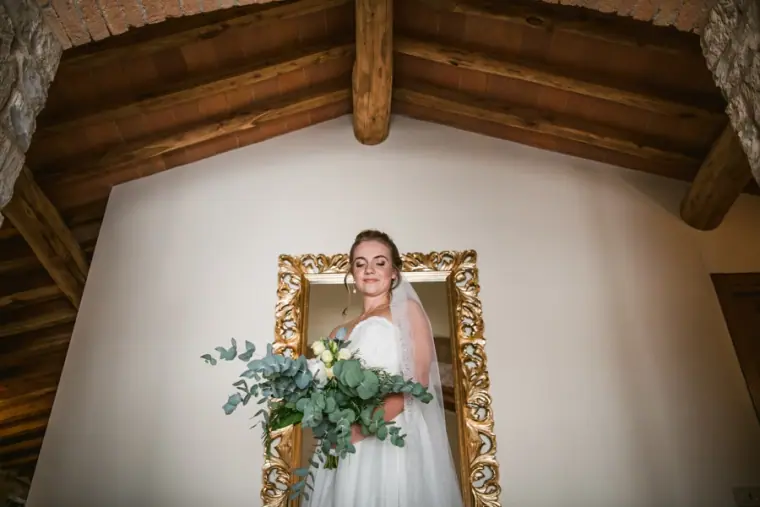 Bride holding a eucalyptus bouquet framed in a gilded mirror under a wooden beamed ceiling