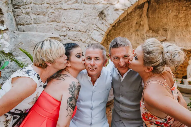 Andy and Andrew being kissed on the cheek by friends in front of a stone wall