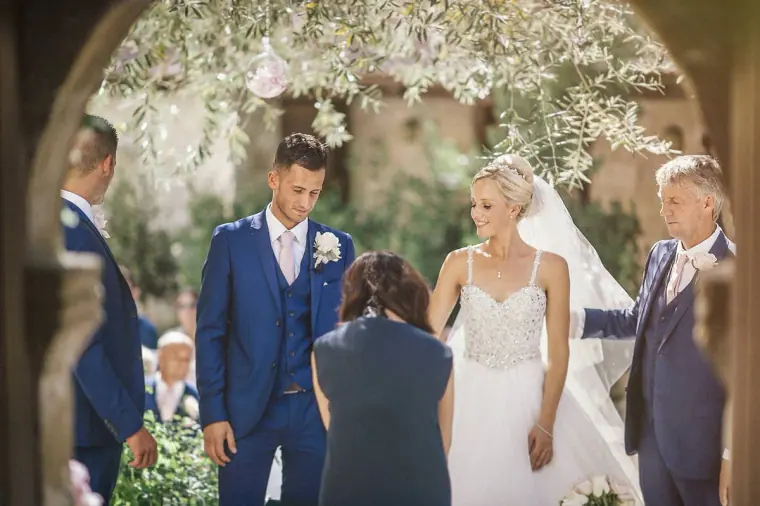 Couple standing before the officiant, framed by a stone arch at Ayii Anargyri