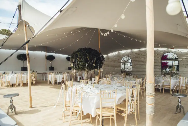 Reception tables set beneath a stretch tent with festoon lights and greenery