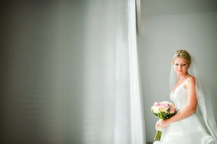 Charlotte in her gown and veil holding her bouquet beside a sheer-curtained window