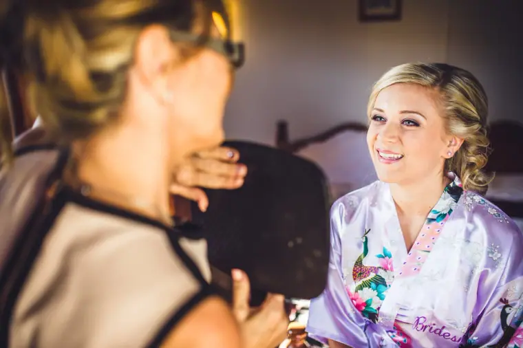 Bride in a floral "Bride" robe smiling at her make-up artist