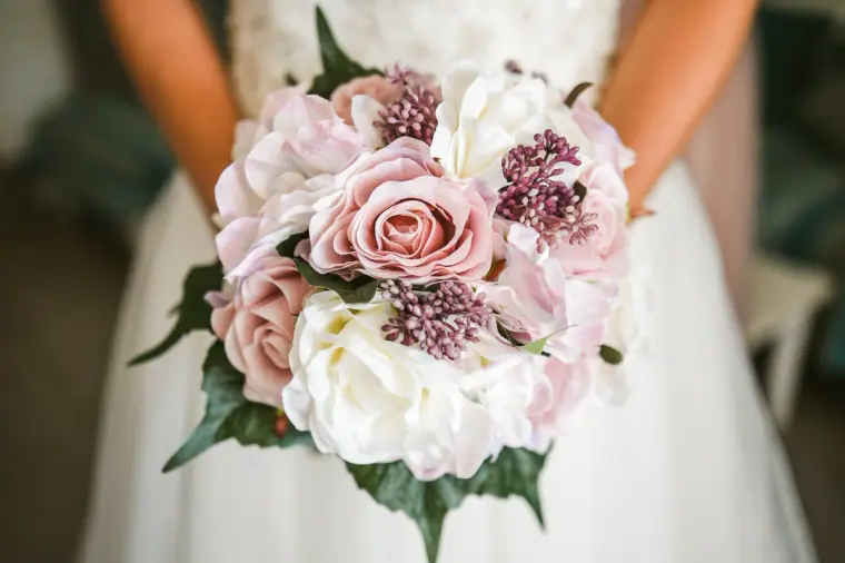 Bride's bouquet of pink roses, white peonies and lilac flowers held against her gown