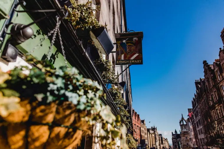 Hanging flower baskets and a shop sign on a sunlit Old Town street, Edinburgh