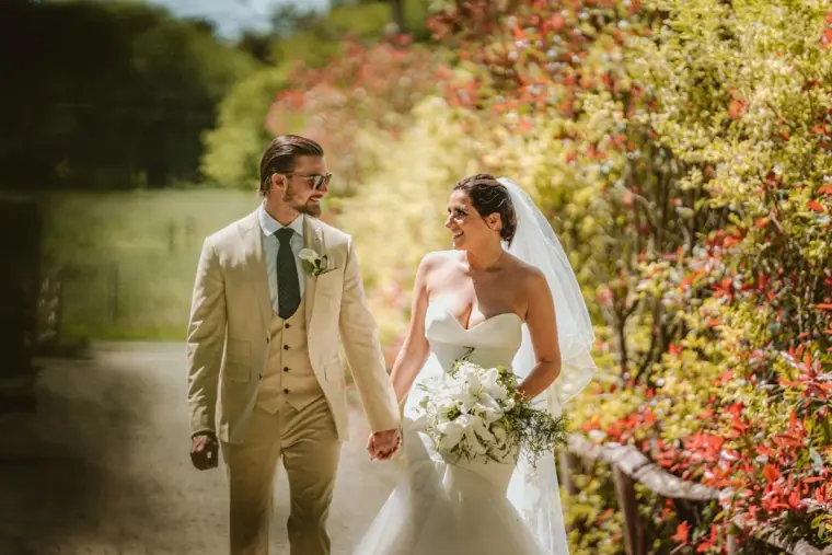 Lauren and Josh walking past a flowering hedge in soft sunlight
