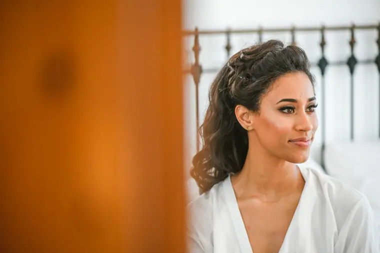 Bride in a white robe gazing out of the bedroom window, hair styled in soft curls