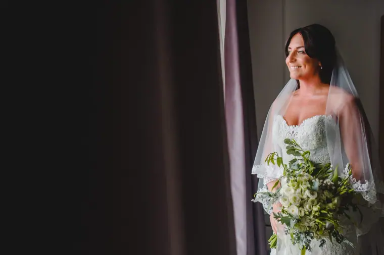 Bride in her veil holding a bouquet in soft window light against a dark curtain