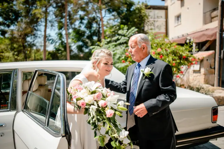 Bride stepping out of a vintage white car with her father, holding a cascading bouquet