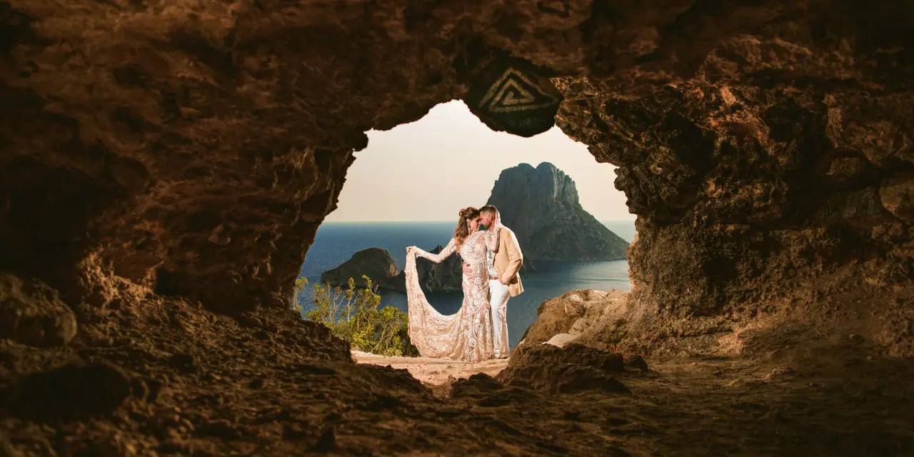 Couple framed by a cave opening with a rocky island rising from the sea beyond