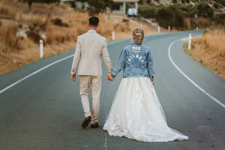 Bride in a denim jacket walking hand in hand with the groom down a winding road