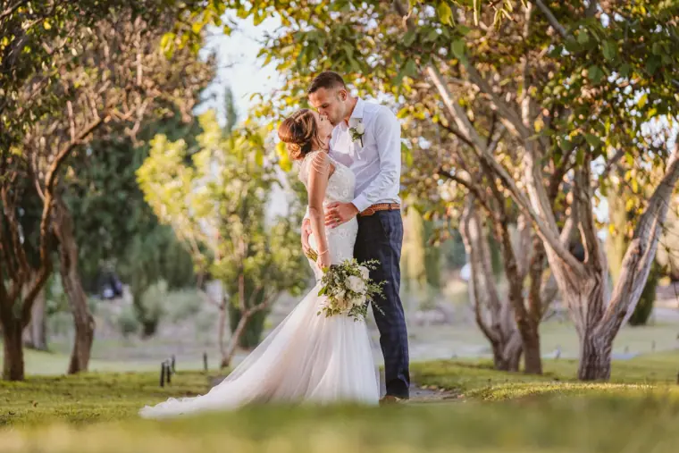 Bride and groom kissing on the lawn under arching olive trees